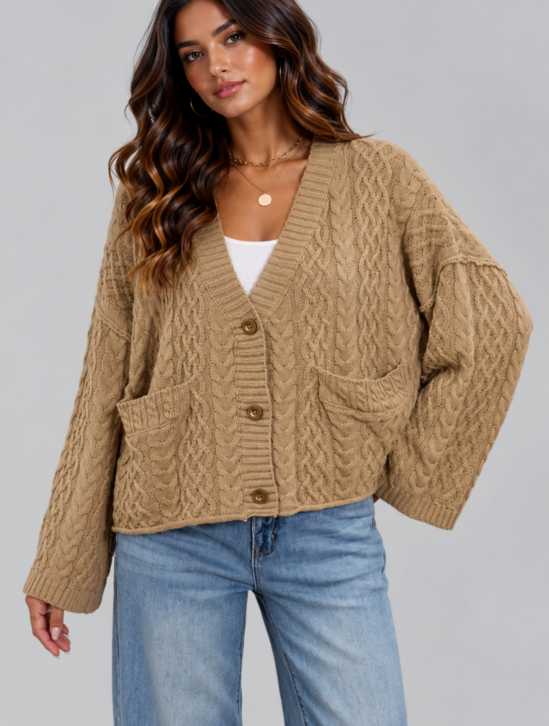 A woman wearing a beige cable-knit cardigan with brown buttons, styled with a white top and blue jeans against a solid gray studio background.