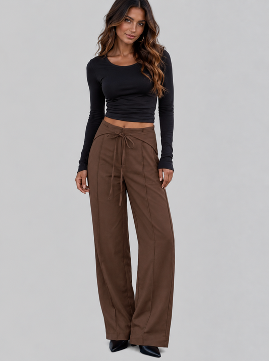 A woman modeling a black long-sleeve crop top and brown wide-leg drawstring pants against a solid gray studio background.
