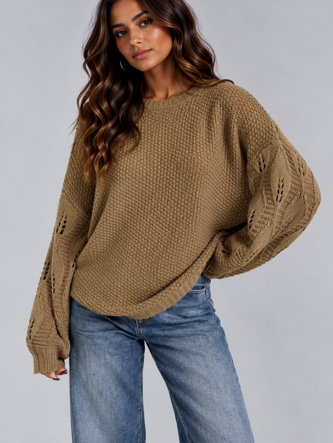 A woman wearing a chunky knit brown sweater with balloon sleeves and blue jeans, photographed against a plain light gray studio background.