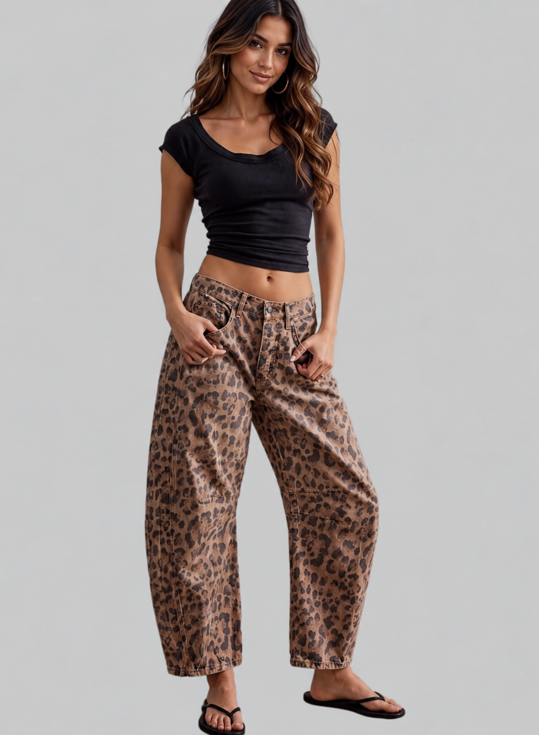 A woman modeling a pair of wide-leg, relaxed-fit pants with a brown and black leopard print pattern, paired with a black crop top and black sandals, against a solid light gray background.