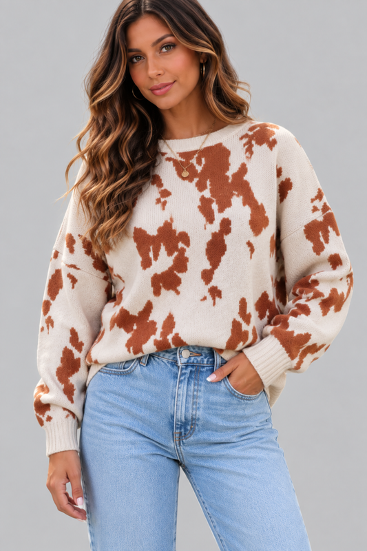 A woman wearing a cream-colored knit sweater with a rust-brown tie-dye pattern, paired with light-wash blue jeans, against a solid gray studio background.