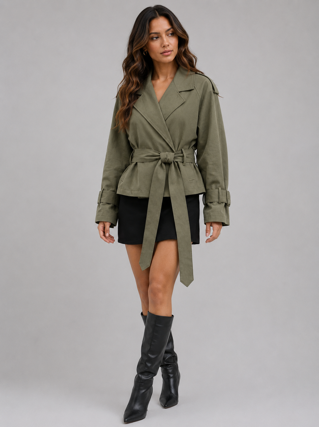 A woman modeling an olive green belted wrap jacket with a black mini skirt and black knee-high boots, standing against a solid gray studio background.