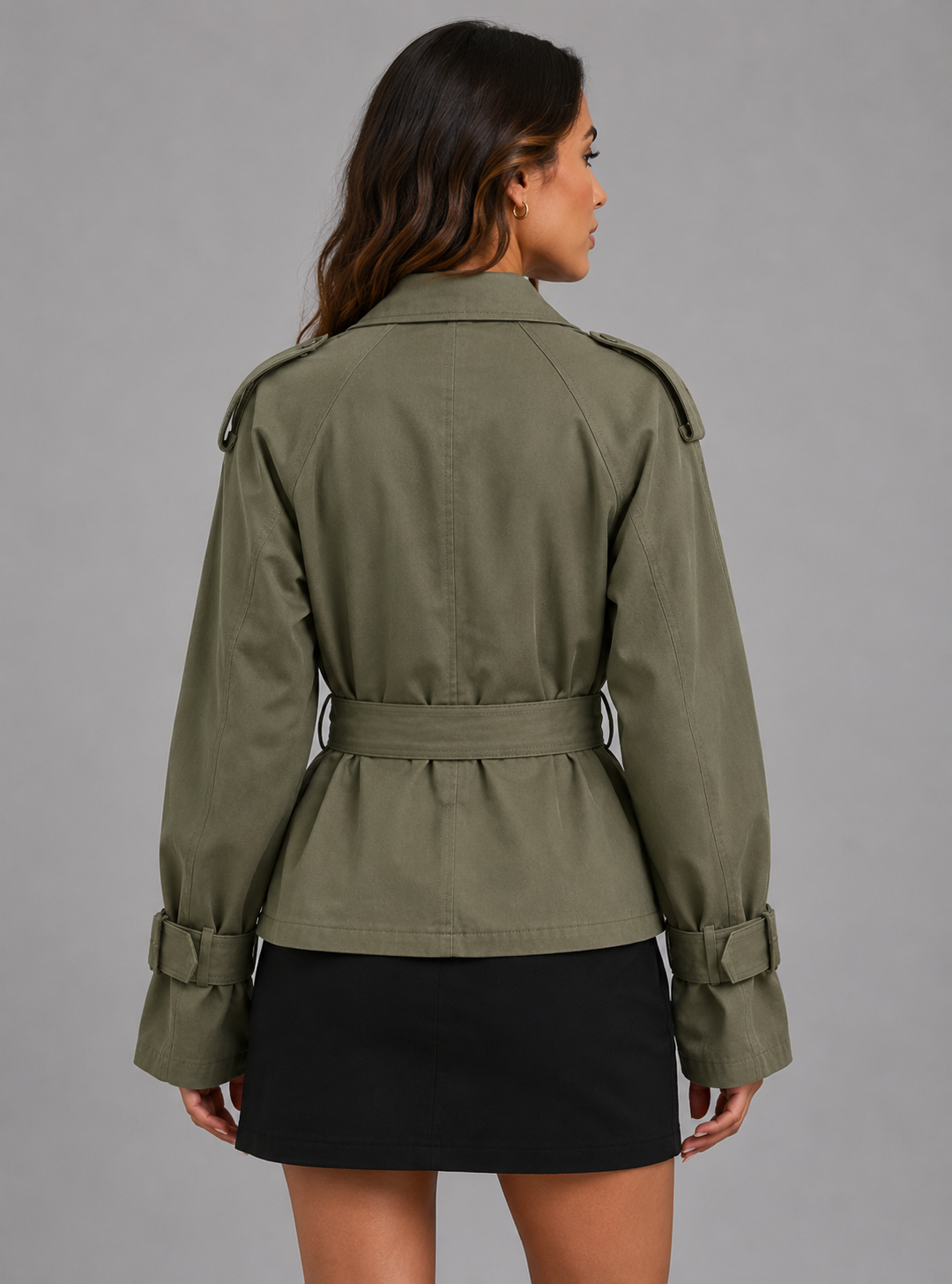 A woman shown from the back wearing an olive green belted jacket and a black skirt against a plain gray studio background.