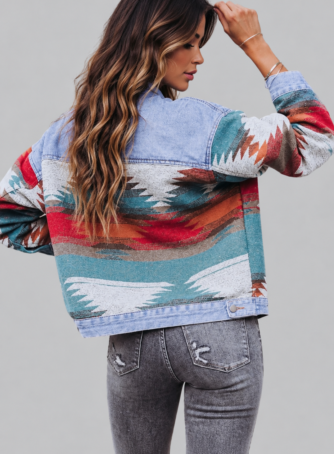 A woman shown from the back wearing a long-sleeve denim jacket with a colorful Southwestern-style pattern in red, teal, white, and orange, paired with gray jeans, against a solid gray background.
