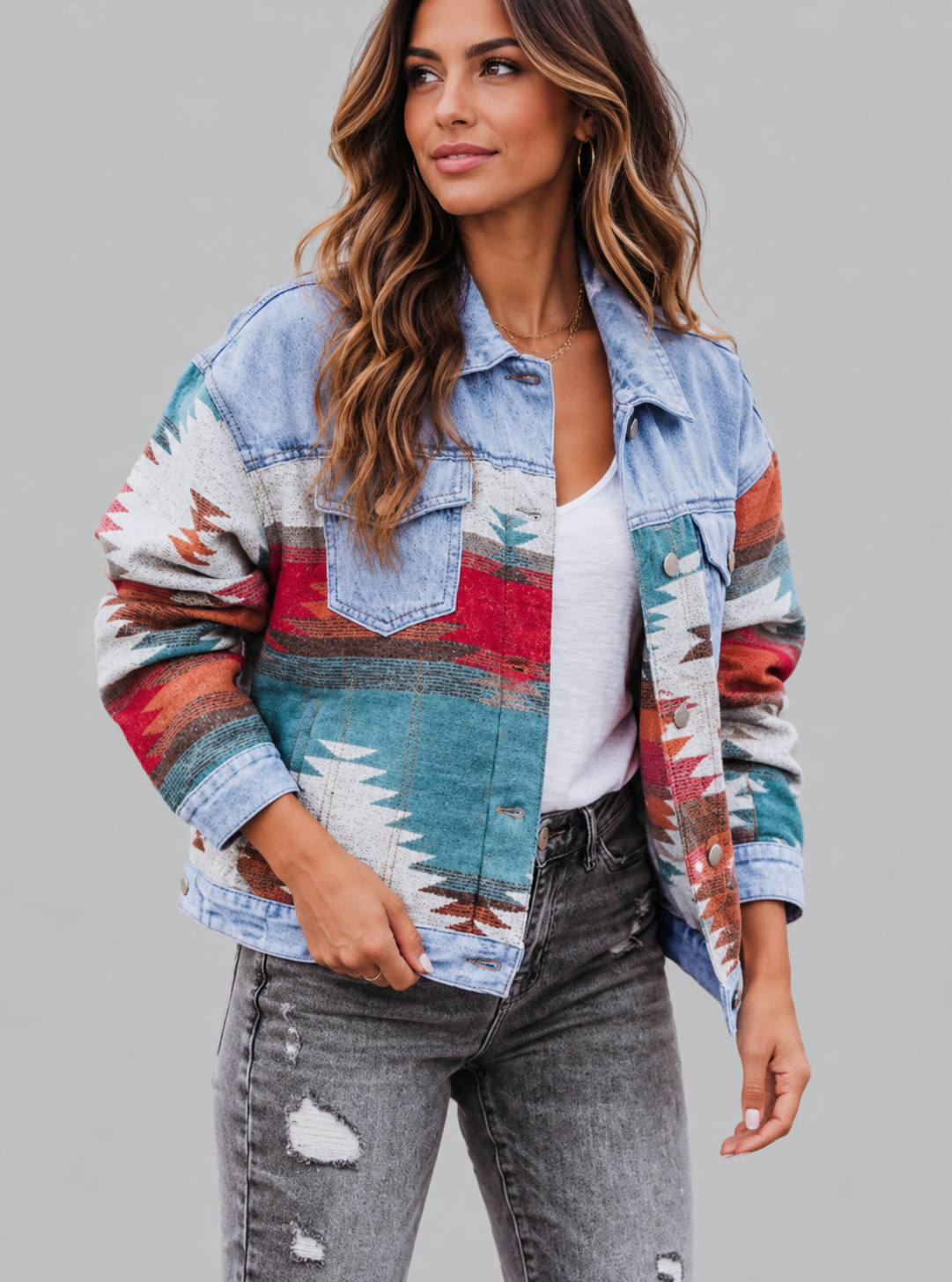 A woman wearing a light blue denim jacket with colorful Southwestern-style geometric and mountain pattern panels, styled with a white top and distressed gray jeans against a solid gray studio background.