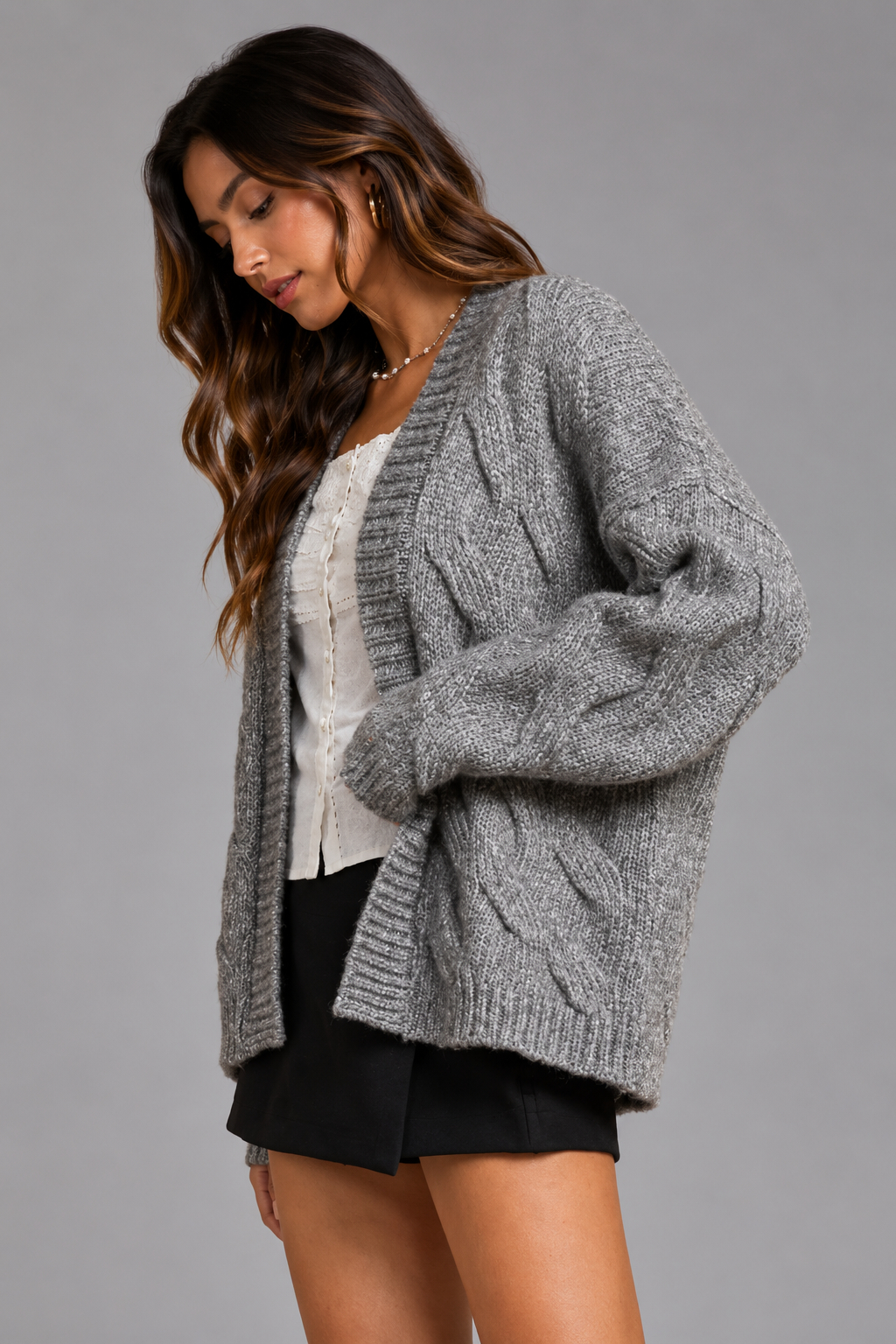 A woman wearing a chunky gray cable-knit cardigan over a white top and black shorts, photographed against a solid gray studio background.
