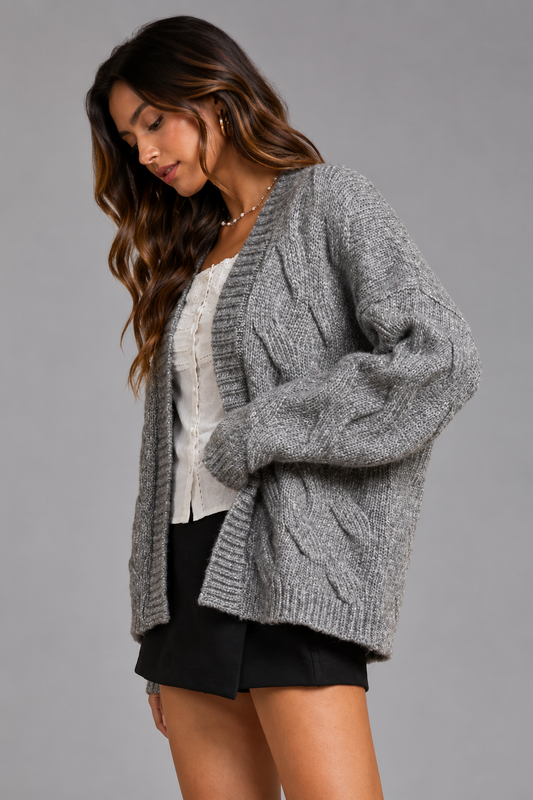 A woman wearing a chunky gray cable-knit cardigan over a white top and black shorts, photographed against a solid gray studio background.