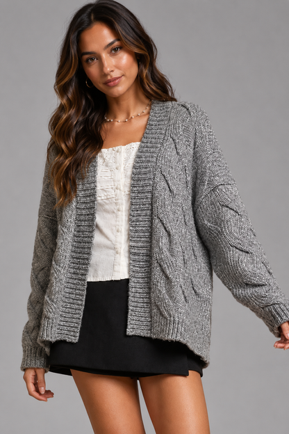 A woman modeling a gray cable-knit open-front cardigan over a white top and black skirt, photographed against a plain gray studio background.