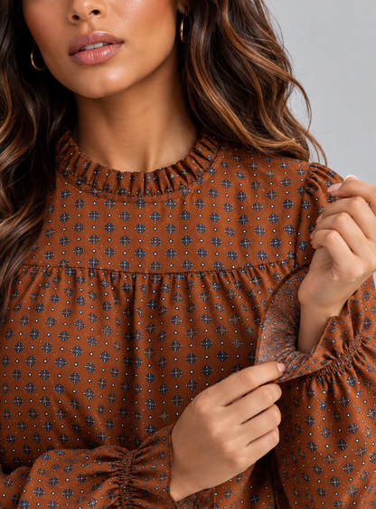 A close-up of a woman wearing a brown long-sleeve blouse with a small geometric print, featuring a ruffled neckline and gathered sleeves.