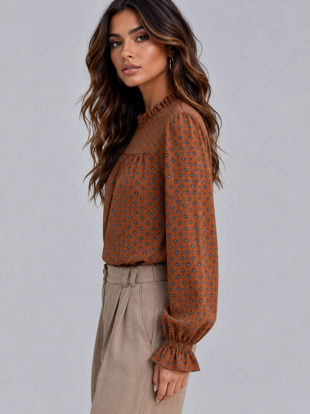 A woman wearing a rust-orange long-sleeve blouse with a small blue and black geometric pattern, paired with beige trousers, photographed against a solid gray studio background.