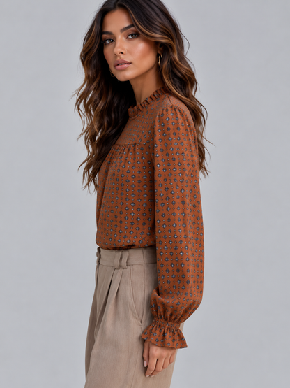 A woman wearing a rust-orange long-sleeve blouse with a small blue and black geometric pattern, paired with beige trousers, photographed against a solid gray studio background.