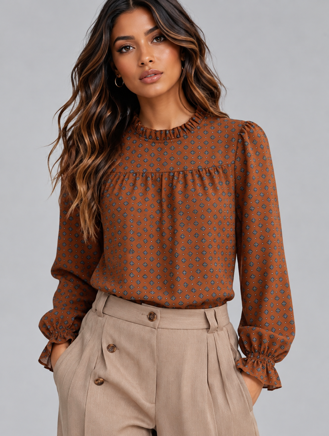 A woman wearing a rust-brown long-sleeve blouse with a small geometric pattern and pleated details, paired with beige high-waisted trousers, against a solid gray studio background.