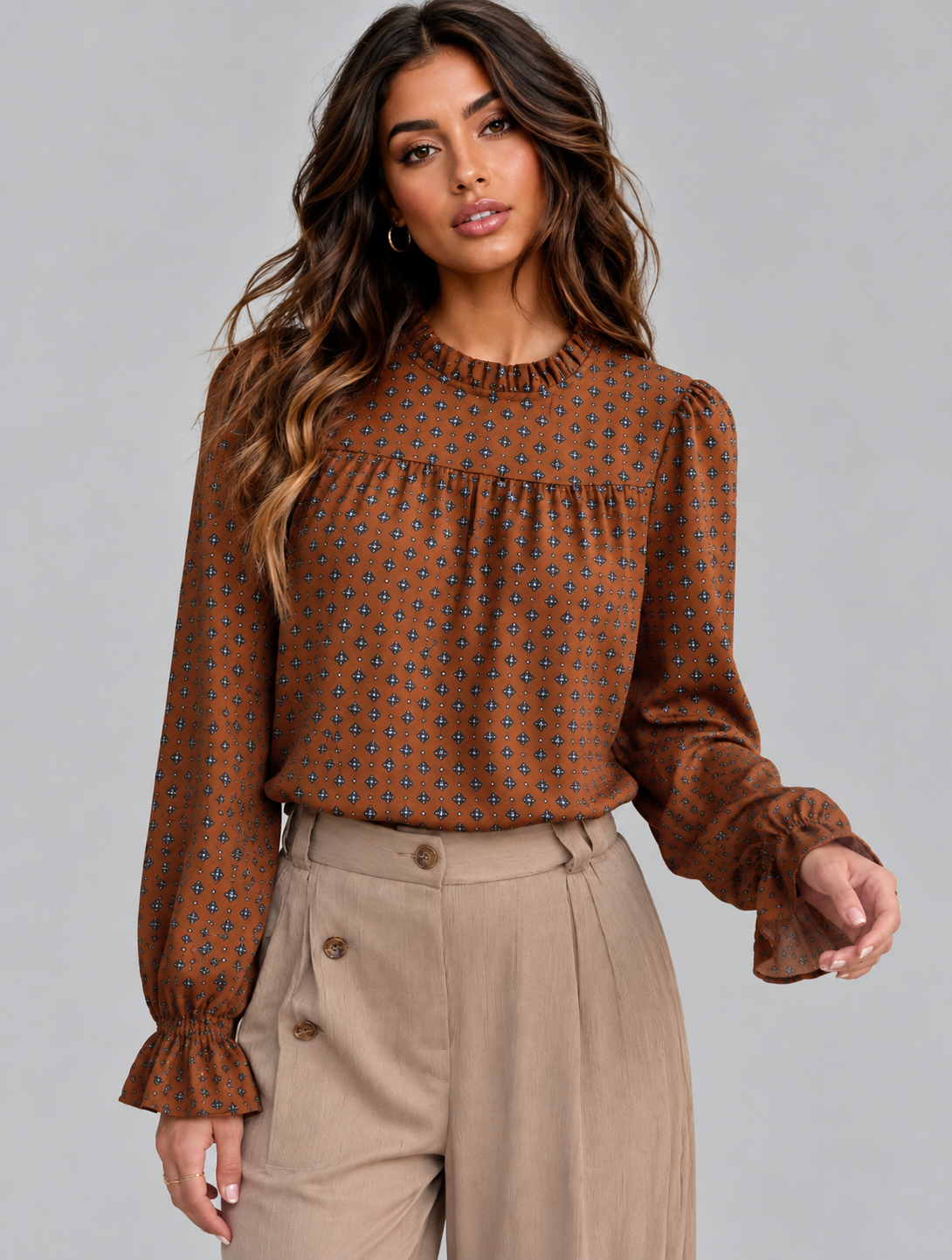 A woman modeling a brown long-sleeve blouse with a small geometric pattern, paired with beige high-waisted trousers, against a solid gray studio background.
