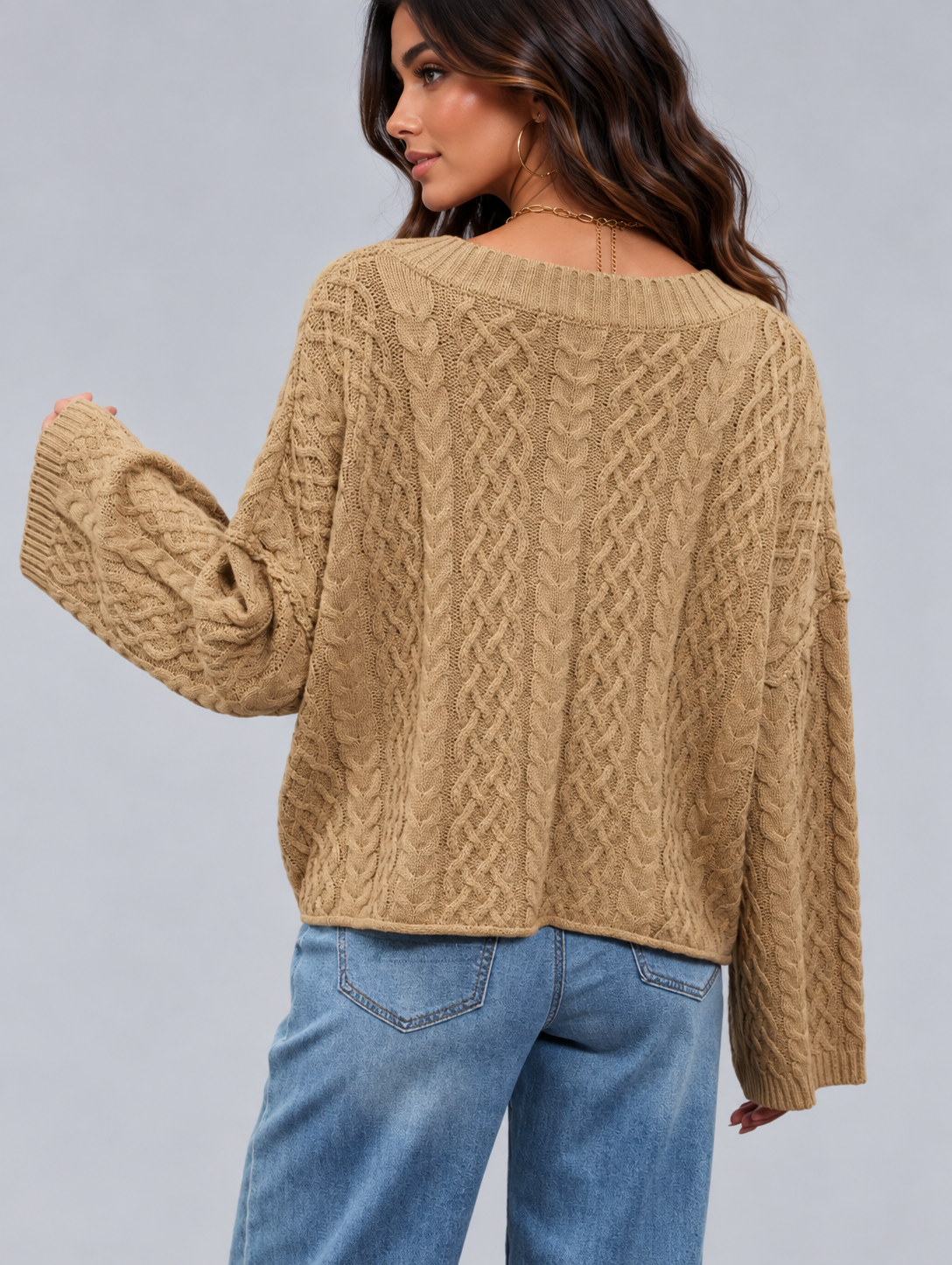 A woman wearing a beige cable-knit sweater with wide sleeves, shown from the back against a solid gray studio background.