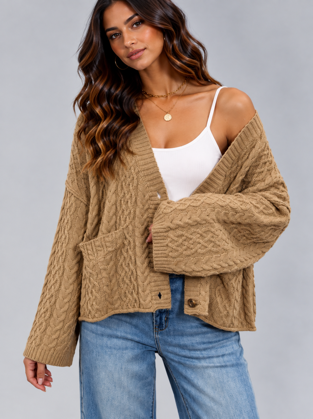 A woman wearing a tan cable-knit cardigan over a white camisole and blue jeans, photographed against a plain light gray studio background.