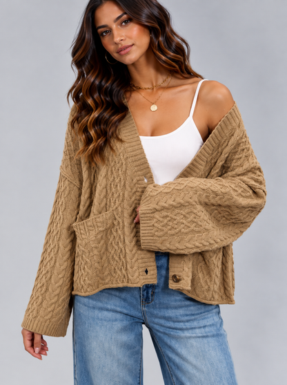 A woman wearing a tan cable-knit cardigan over a white camisole and blue jeans, photographed against a plain light gray studio background.
