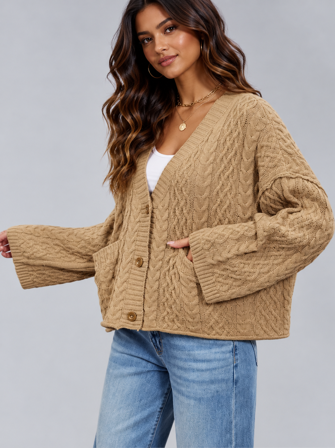 A woman wearing a cropped, cable-knit cardigan in a warm camel brown color, styled with a white top and blue jeans against a solid gray studio background.