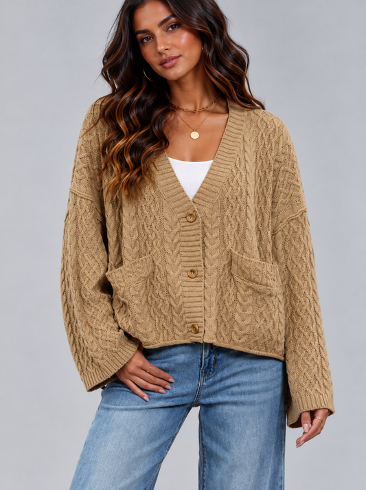 A woman wearing a camel-colored cable-knit cardigan with front buttons and patch pockets, styled over a white top and blue jeans, against a solid gray studio background.