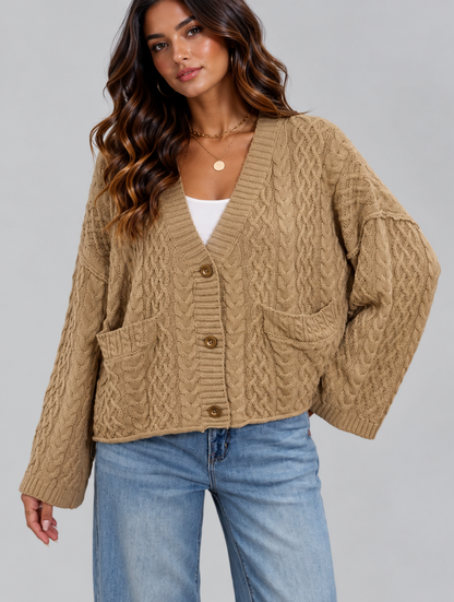 A woman wearing a beige cable-knit cardigan with brown buttons, styled with a white top and blue jeans against a solid gray studio background.
