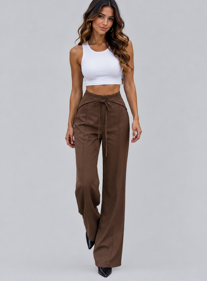 A woman modeling a pair of high-waisted, wide-leg brown pants with a drawstring waist, paired with a white crop top, against a solid light gray background.