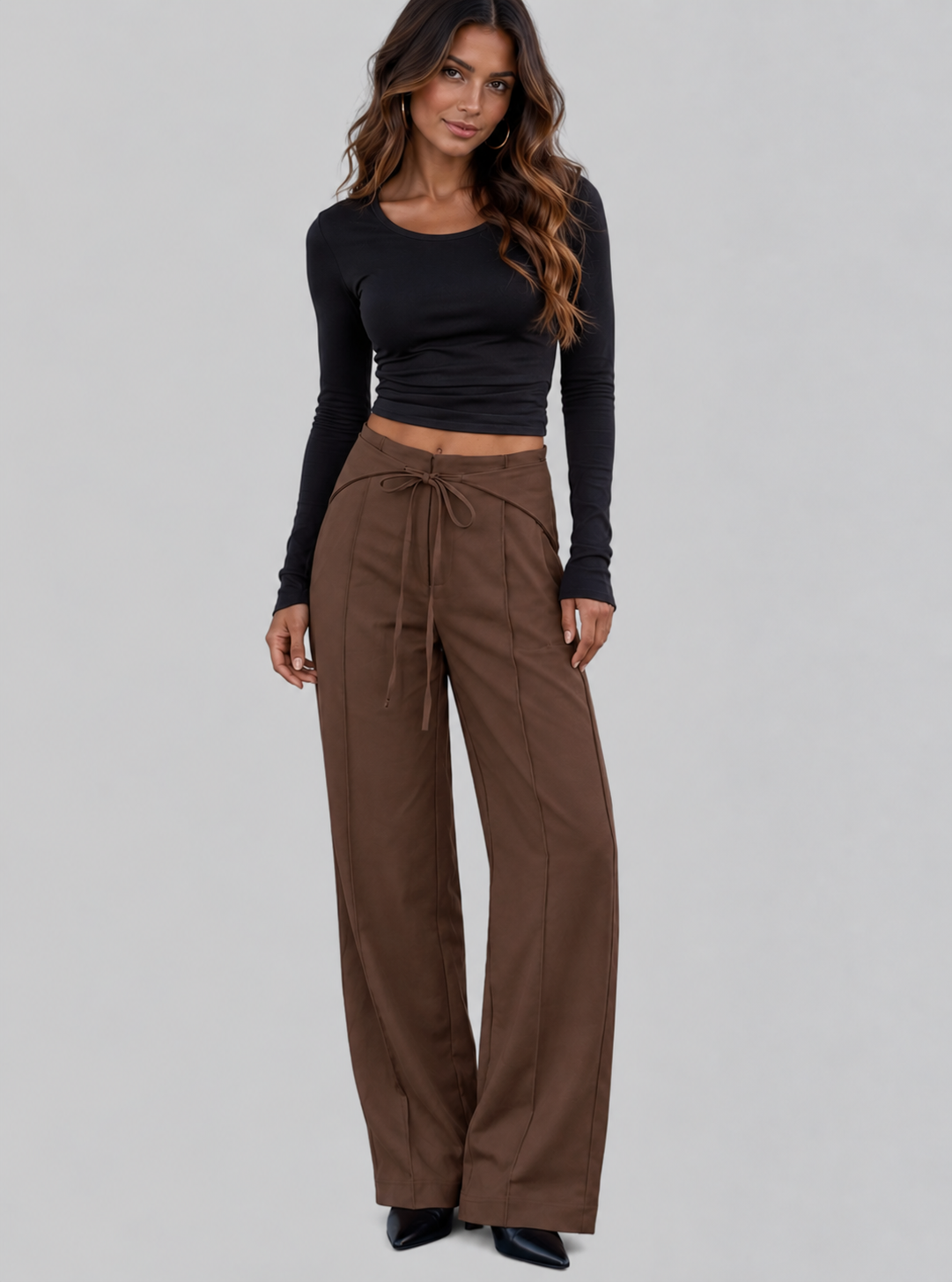 A woman modeling a black long-sleeve crop top and brown wide-leg drawstring pants against a solid gray studio background.