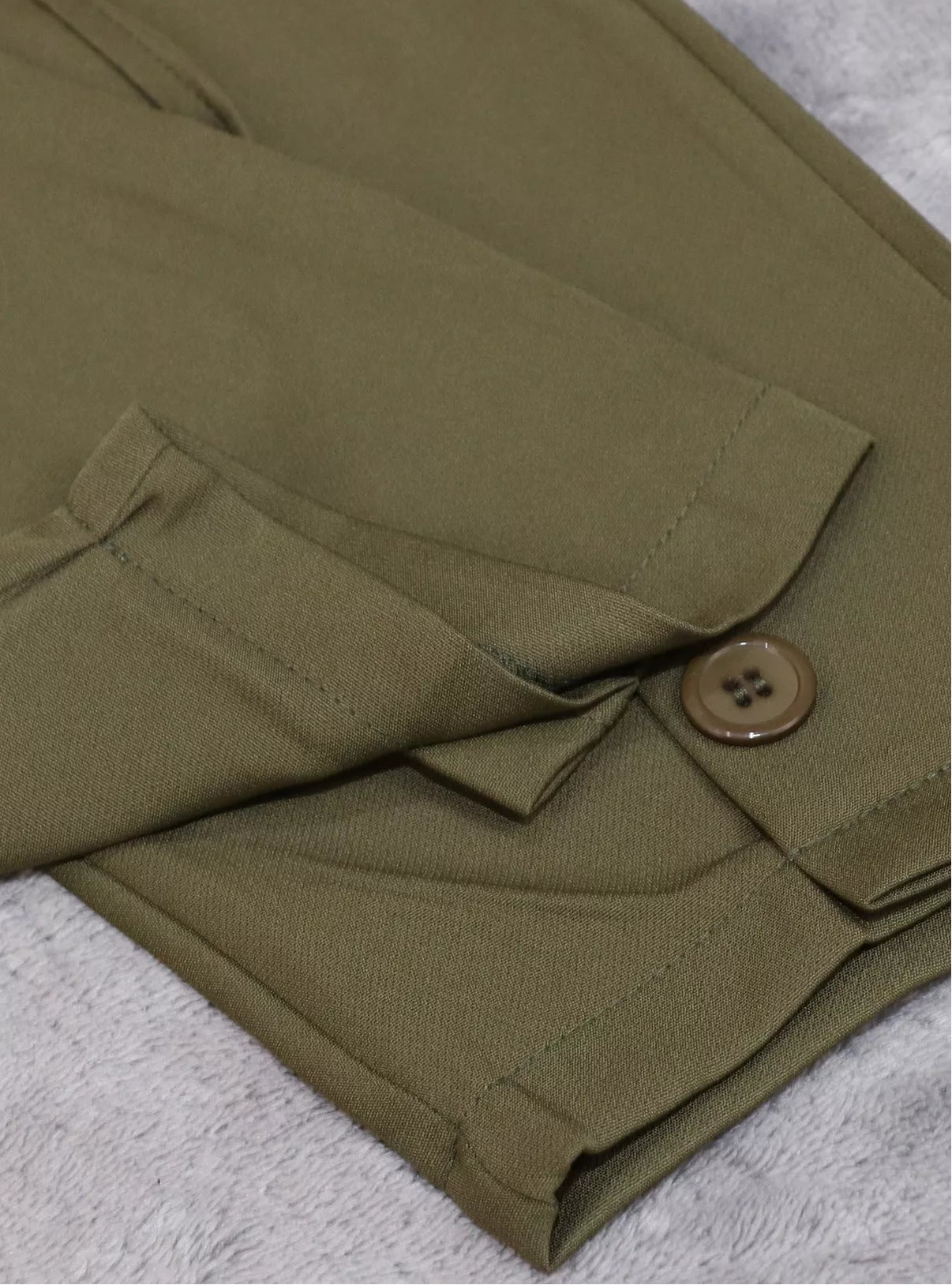 Close-up of olive green trousers folded on a textured light gray surface, showing a buttoned flap pocket and fabric texture.