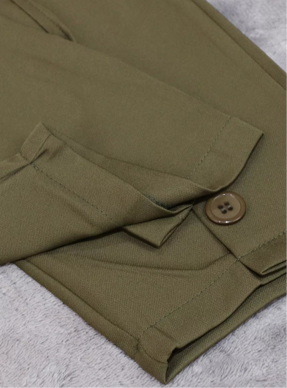 Close-up of olive green trousers folded on a textured light gray surface, showing a buttoned flap pocket and fabric texture.