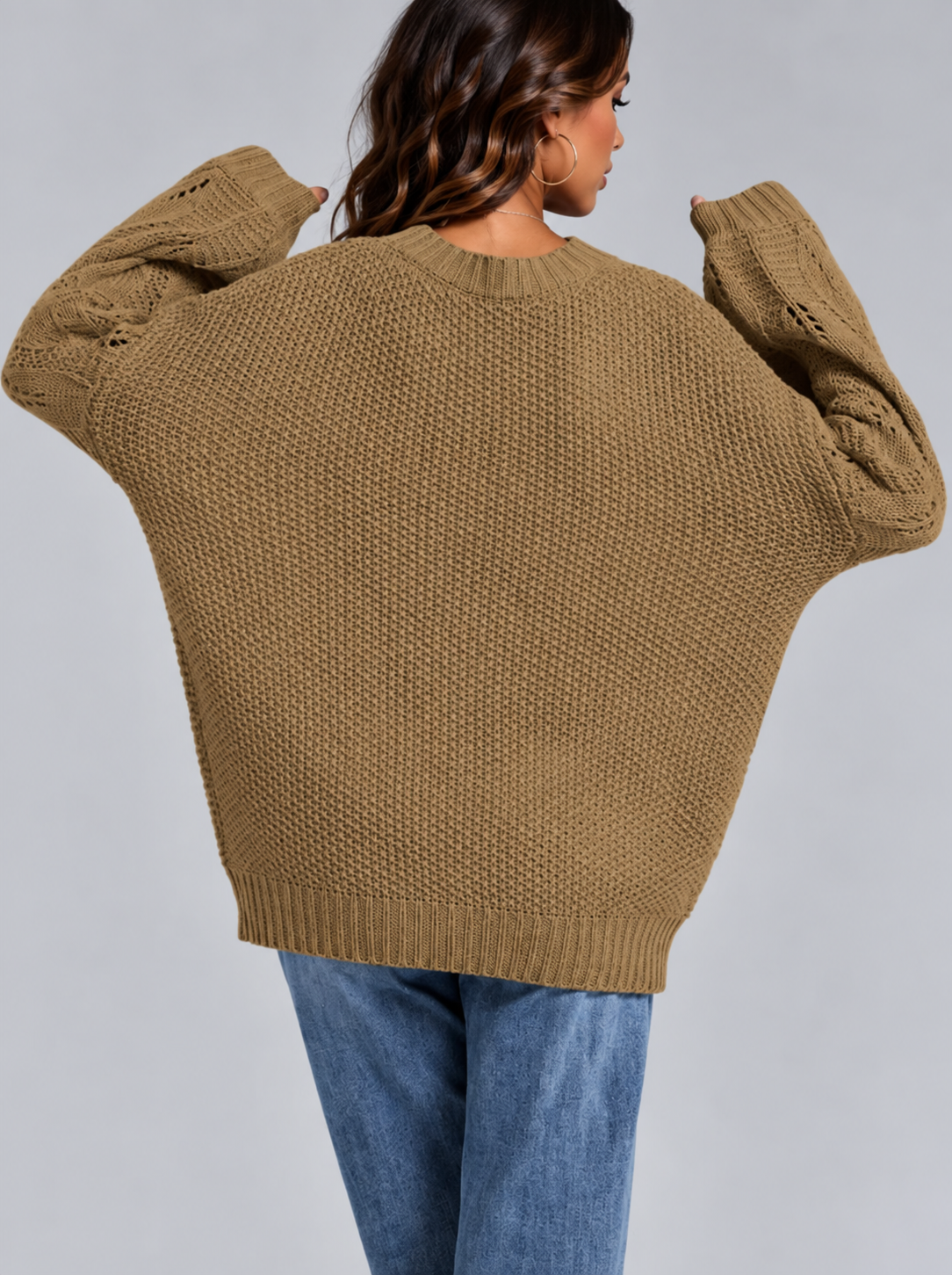 A woman wearing an oversized chunky knit sweater in a warm camel brown color, shown from the back against a solid gray studio background.
