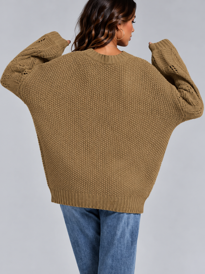 A woman wearing an oversized chunky knit sweater in a warm camel brown color, shown from the back against a solid gray studio background.