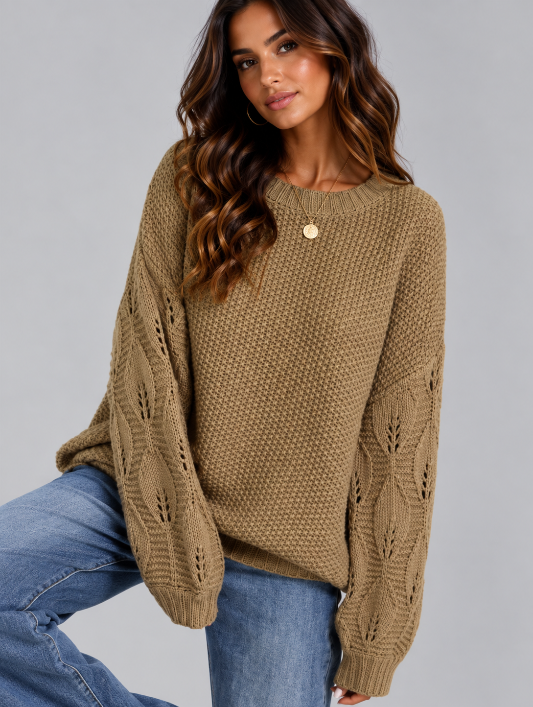 A woman wearing a chunky knit crewneck sweater in a warm camel brown color, paired with blue jeans, posed against a solid gray studio background.