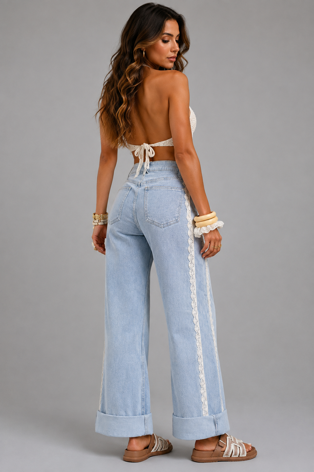 A woman shown from the back wearing light-wash wide-leg jeans with a decorative cream lace detail running down the side, paired with a cream tie-front crop top and sandals, against a solid gray studio background.