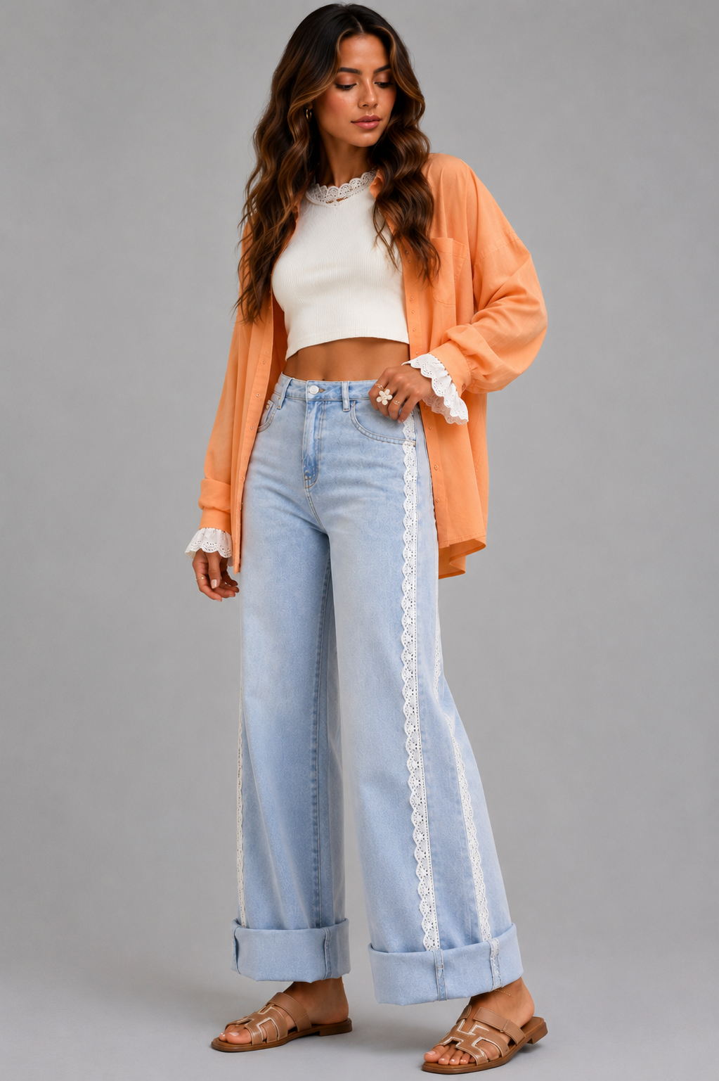 A woman modeling a pair of light-wash wide-leg jeans with white lace side panels, styled with a white crop top, an orange cardigan, and brown sandals against a solid gray studio background.