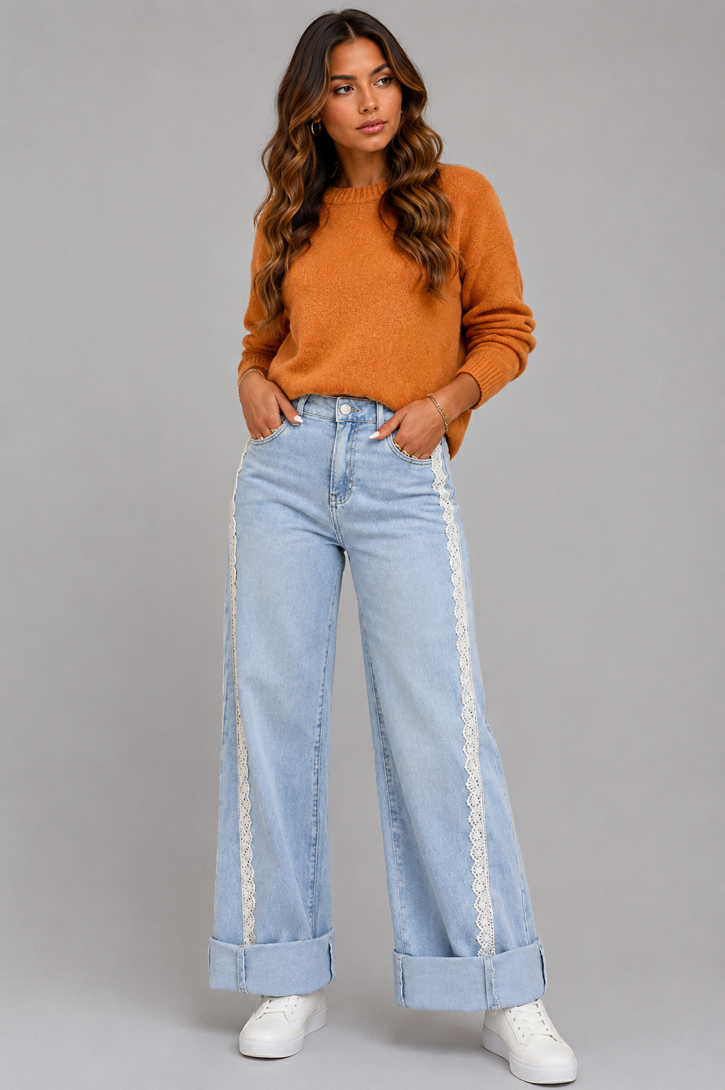 A woman modeling a pair of light-wash wide-leg jeans with white lace side panels, paired with an orange knit sweater and white sneakers, against a solid gray studio background.