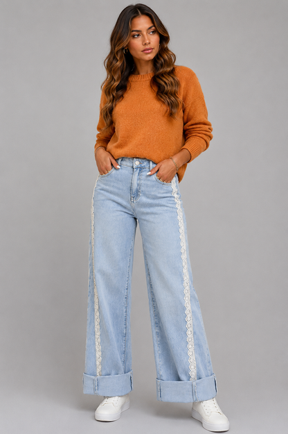 A woman modeling a pair of light-wash wide-leg jeans with white lace side panels, paired with an orange knit sweater and white sneakers, against a solid gray studio background.
