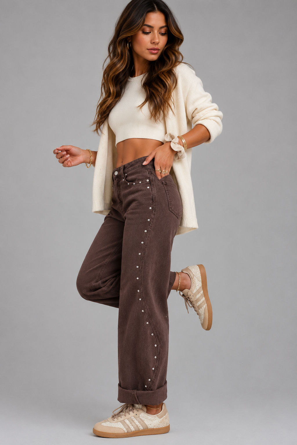 A woman modeling a pair of brown studded wide-leg jeans, styled with a cream crop top, cream cardigan, and beige sneakers, against a solid gray studio background.