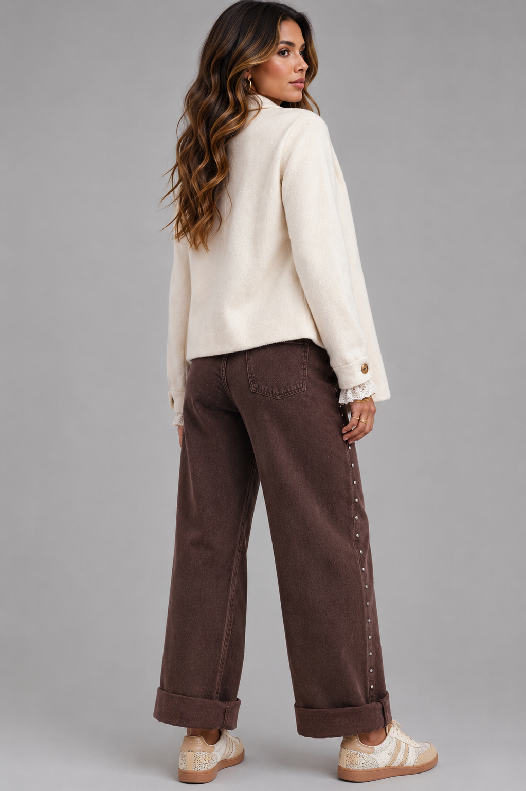 A woman modeling a cream-colored knit cardigan and dark brown wide-leg pants with decorative studs down the side, paired with beige sneakers, against a solid gray studio background.