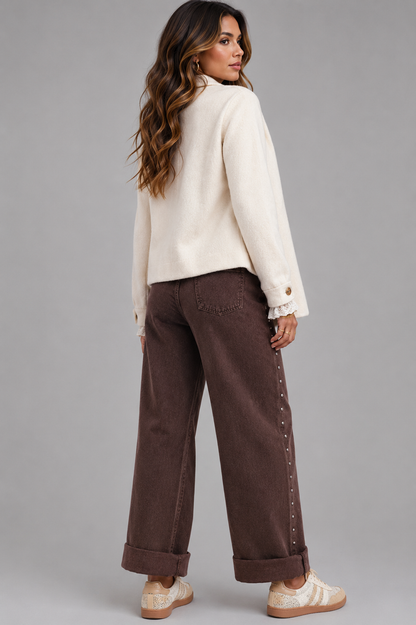 A woman modeling a cream-colored knit cardigan and dark brown wide-leg pants with decorative studs down the side, paired with beige sneakers, against a solid gray studio background.