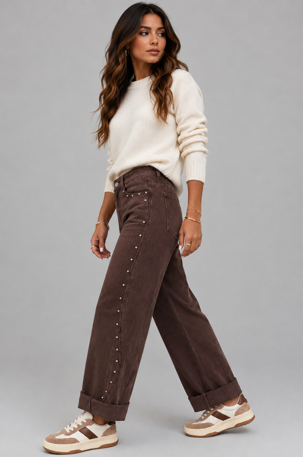 A woman modeling a pair of dark brown wide-leg corduroy pants with silver stud embellishments down the side, paired with a cream knit sweater and beige sneakers, against a solid gray studio background.