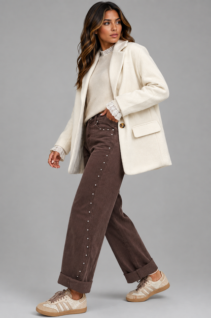 A woman modeling a cream-colored blazer, brown corduroy pants with silver studs, a cream lace-trimmed top, and beige sneakers against a solid gray studio background.