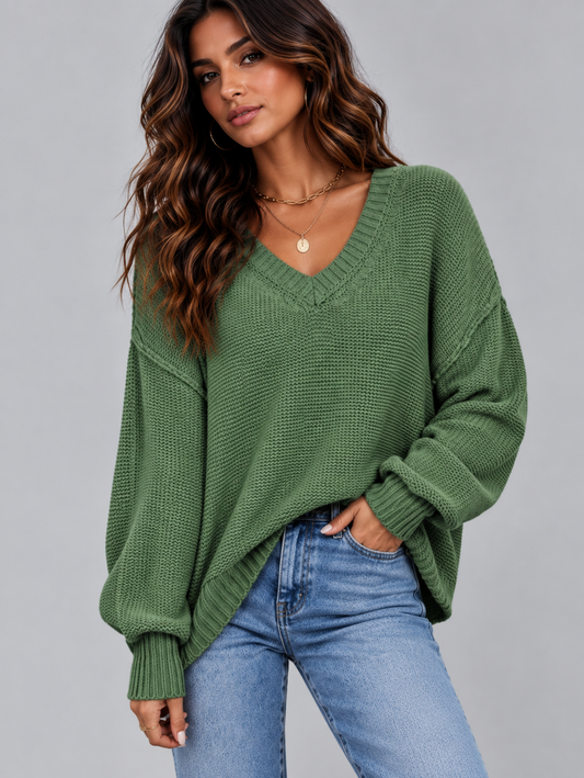 A woman wearing an oversized green ribbed knit sweater with a V-neck and dropped shoulders, paired with light blue jeans, against a solid gray studio background.
