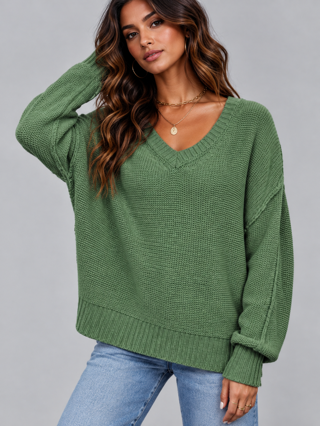 A woman wearing a green ribbed knit V-neck sweater with long sleeves and light blue jeans, posed against a solid gray studio background.