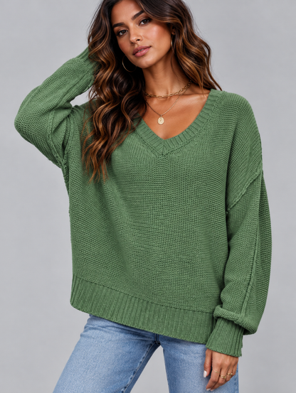 A woman wearing a green ribbed knit V-neck sweater with long sleeves and light blue jeans, posed against a solid gray studio background.