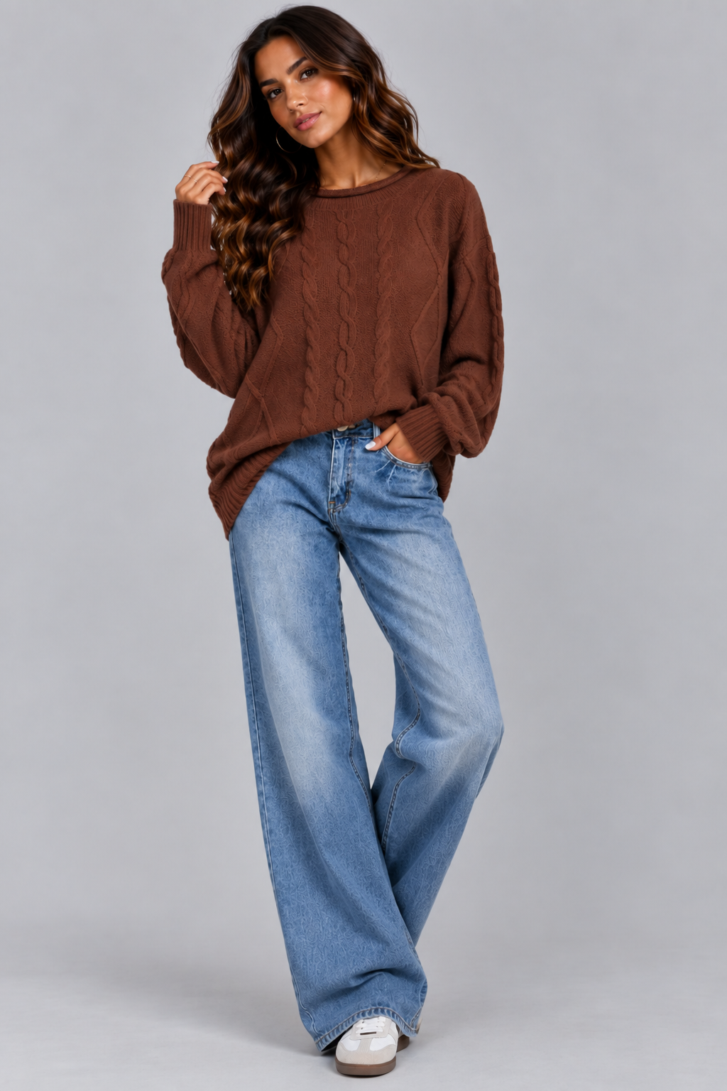 A woman modeling a brown cable-knit crewneck sweater and wide-leg blue jeans against a solid gray studio background.