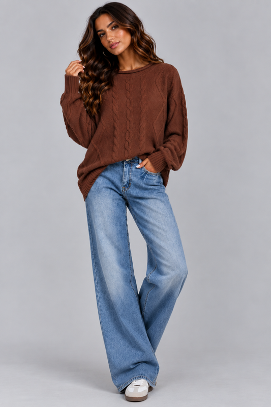 A woman modeling a brown cable-knit crewneck sweater and wide-leg blue jeans against a solid gray studio background.