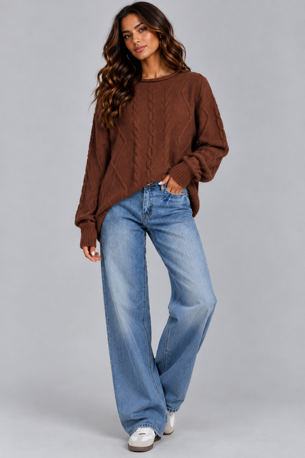 A woman modeling a brown cable-knit crewneck sweater paired with wide-leg blue jeans and white sneakers, photographed against a solid gray studio background.