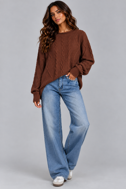 A woman modeling a brown cable-knit crewneck sweater paired with wide-leg blue jeans and white sneakers, photographed against a solid gray studio background.