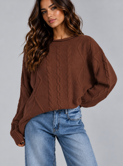A woman wearing a brown cable-knit cropped sweater and light blue jeans, photographed against a plain light gray studio background.