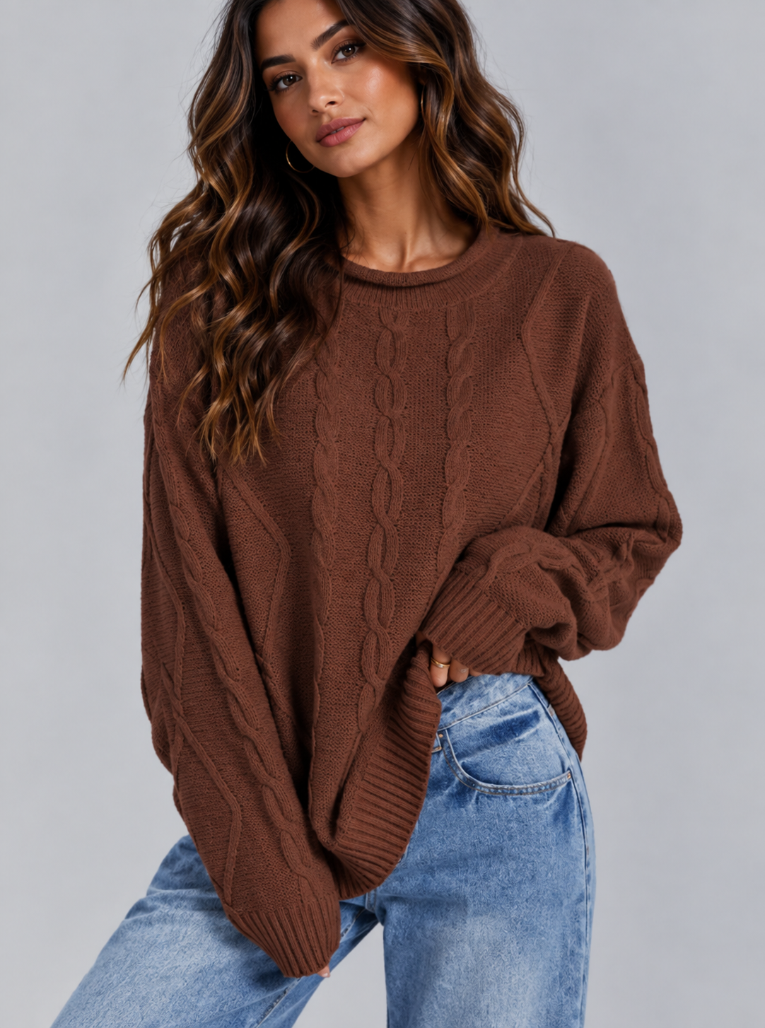 A woman wearing a brown cable-knit crewneck sweater and light blue jeans, posing against a solid gray studio background.