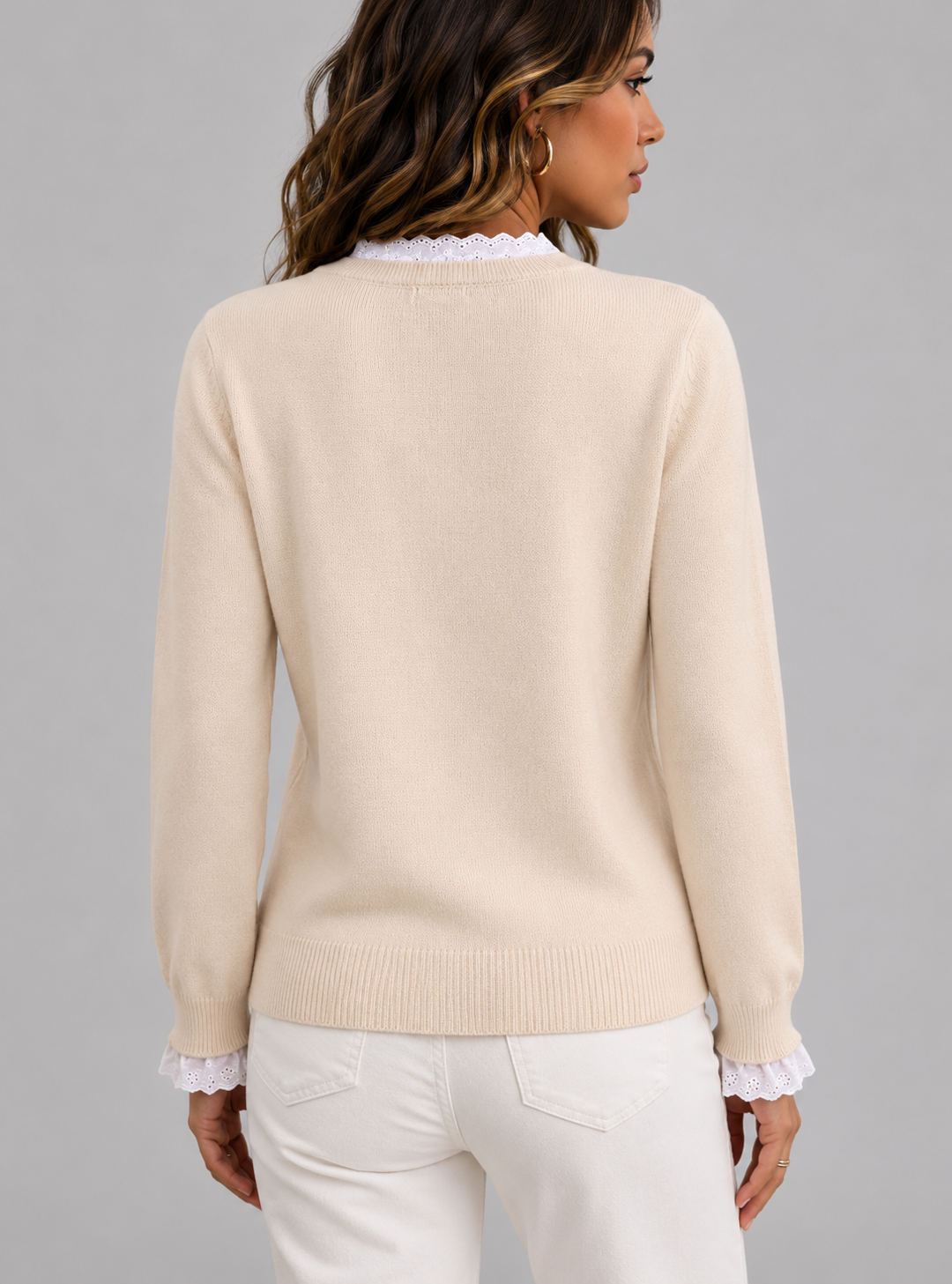 A woman shown from the back wearing a cream-colored knit sweater with white ruffled trim at the crew neck and cuffs, paired with white pants, against a solid gray studio background.
