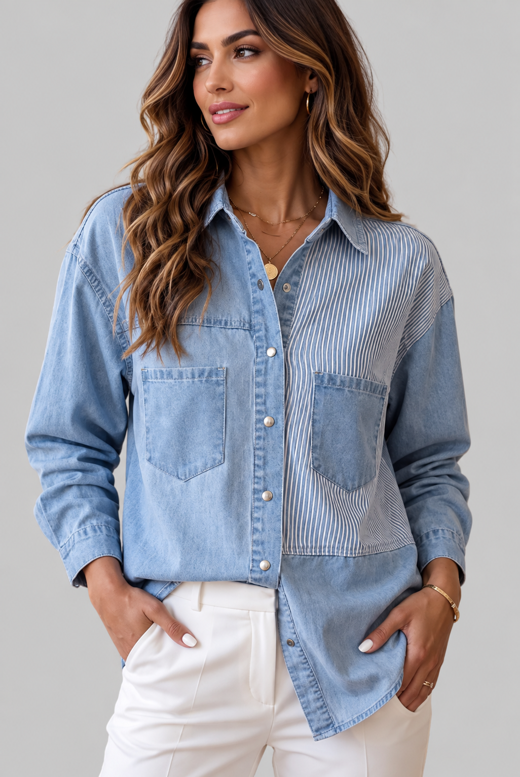 A woman wearing a light blue patchwork button-up shirt with denim and vertically striped panels, paired with white pants, against a solid gray studio background.